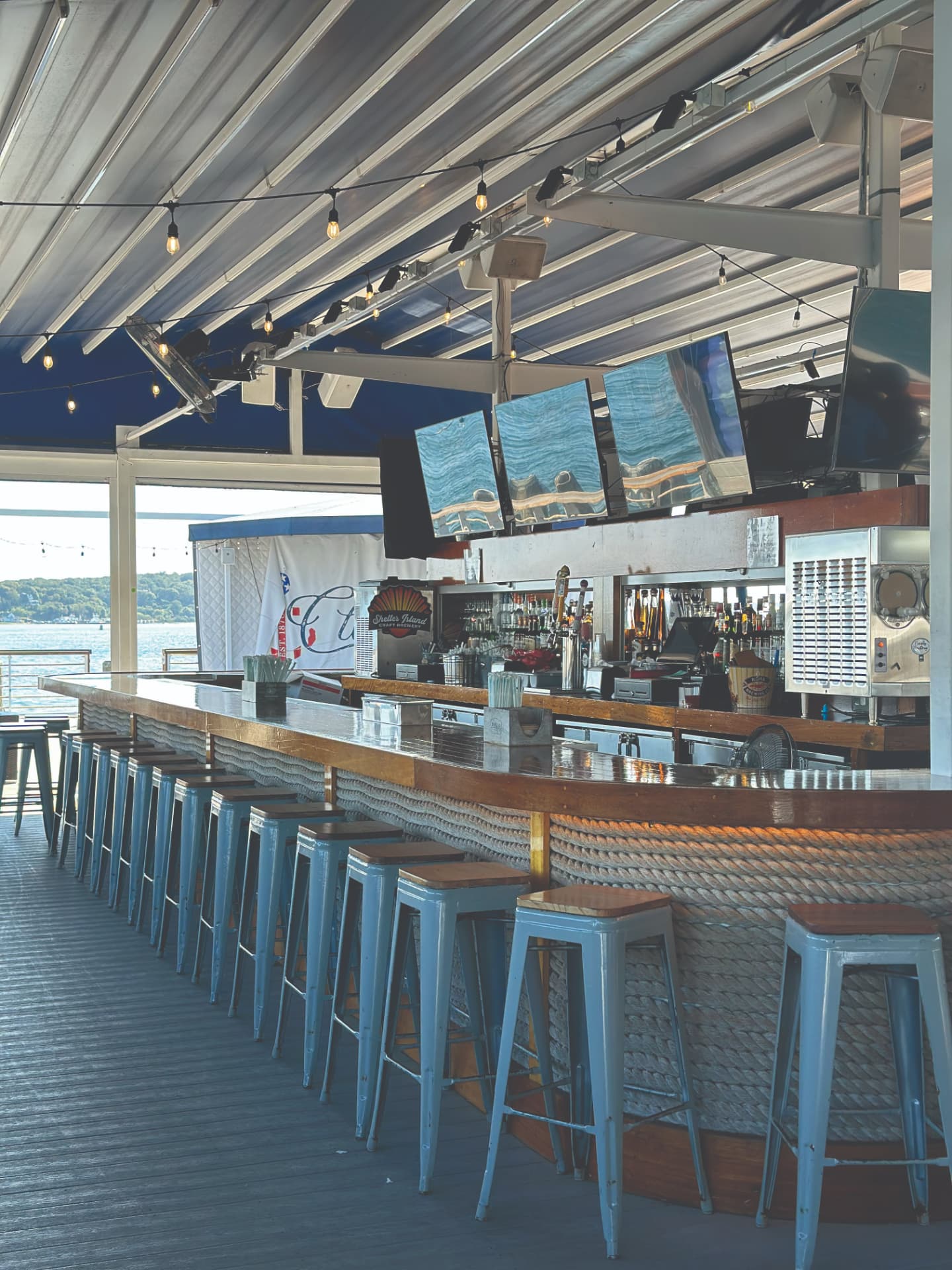 Claudio's waterfront bar with a curved wooden counter, rope-wrapped base, bar stools, string lights, and a view of the marina
